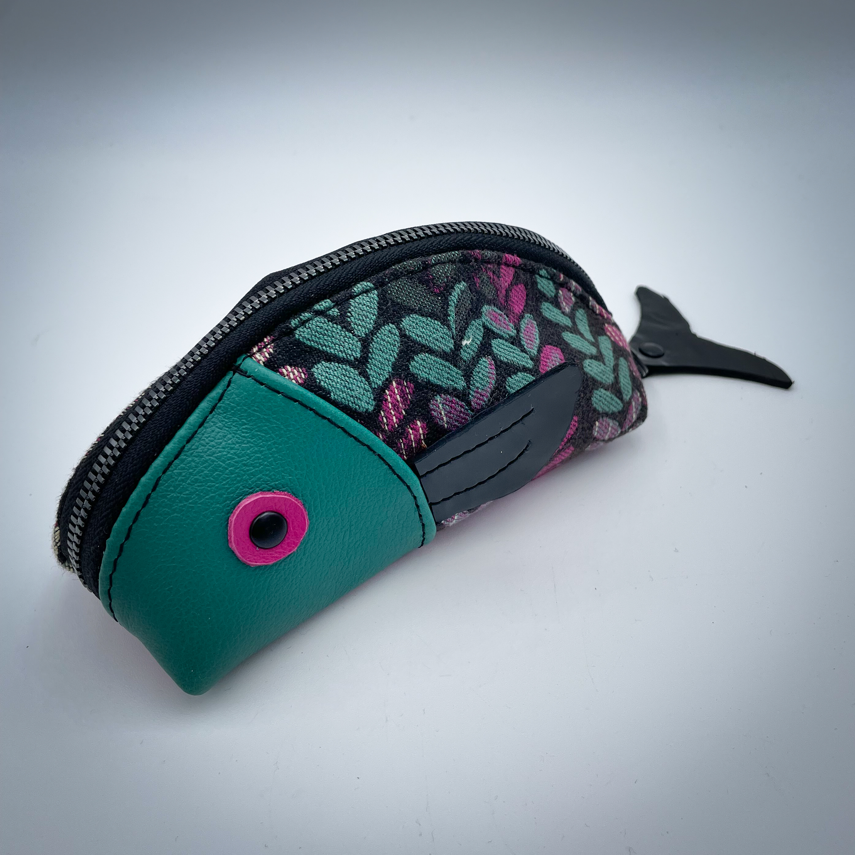 A zippered pouch sewn from sage green leather, black patent leather, fuchsia pink leather, and two scraps of a babywearing wrap with a green, beige, and pink mesh pattern on a black background.